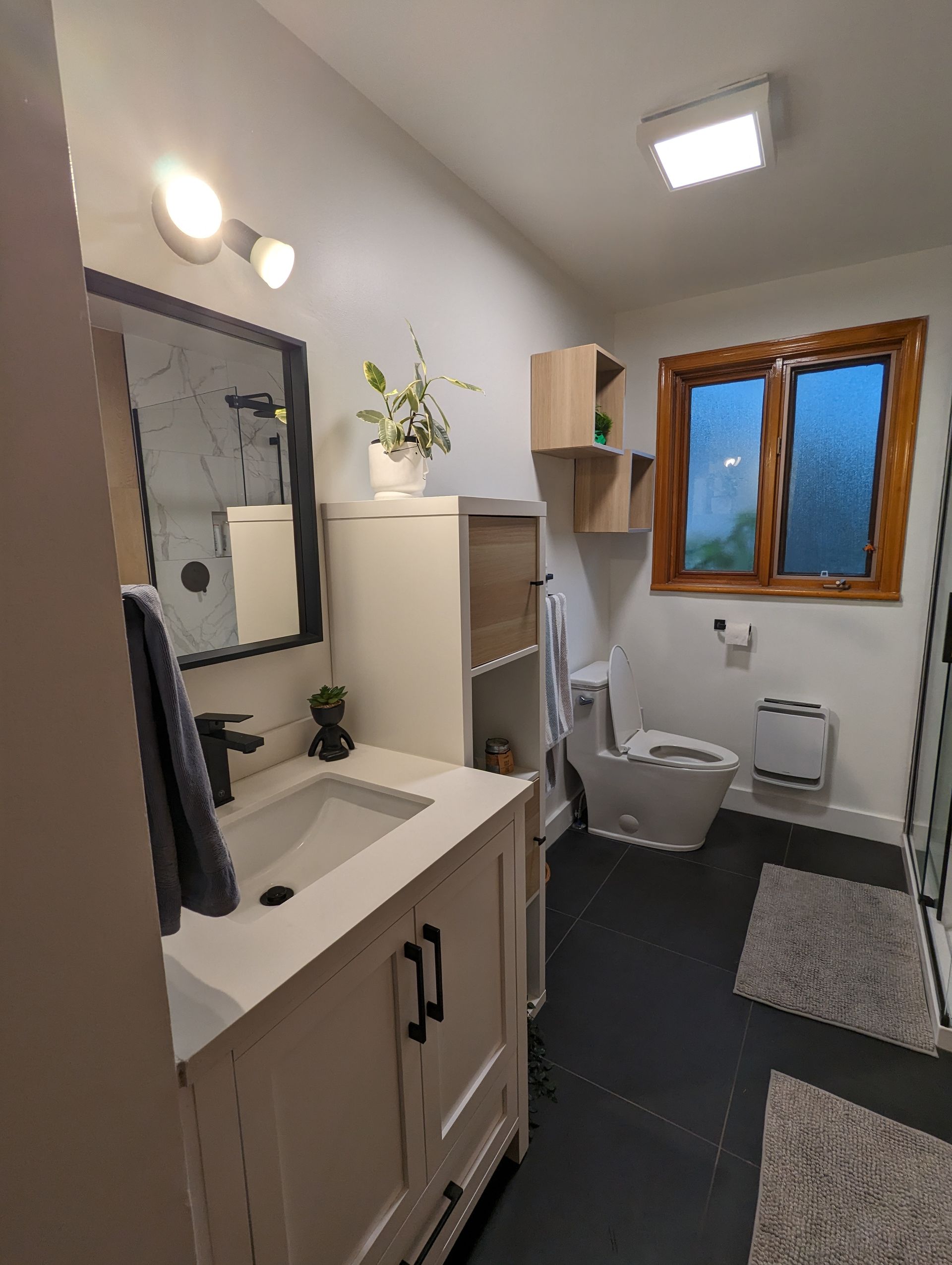Salle de bain avec vanité blanche, sol noir, fenêtre et toilettes.