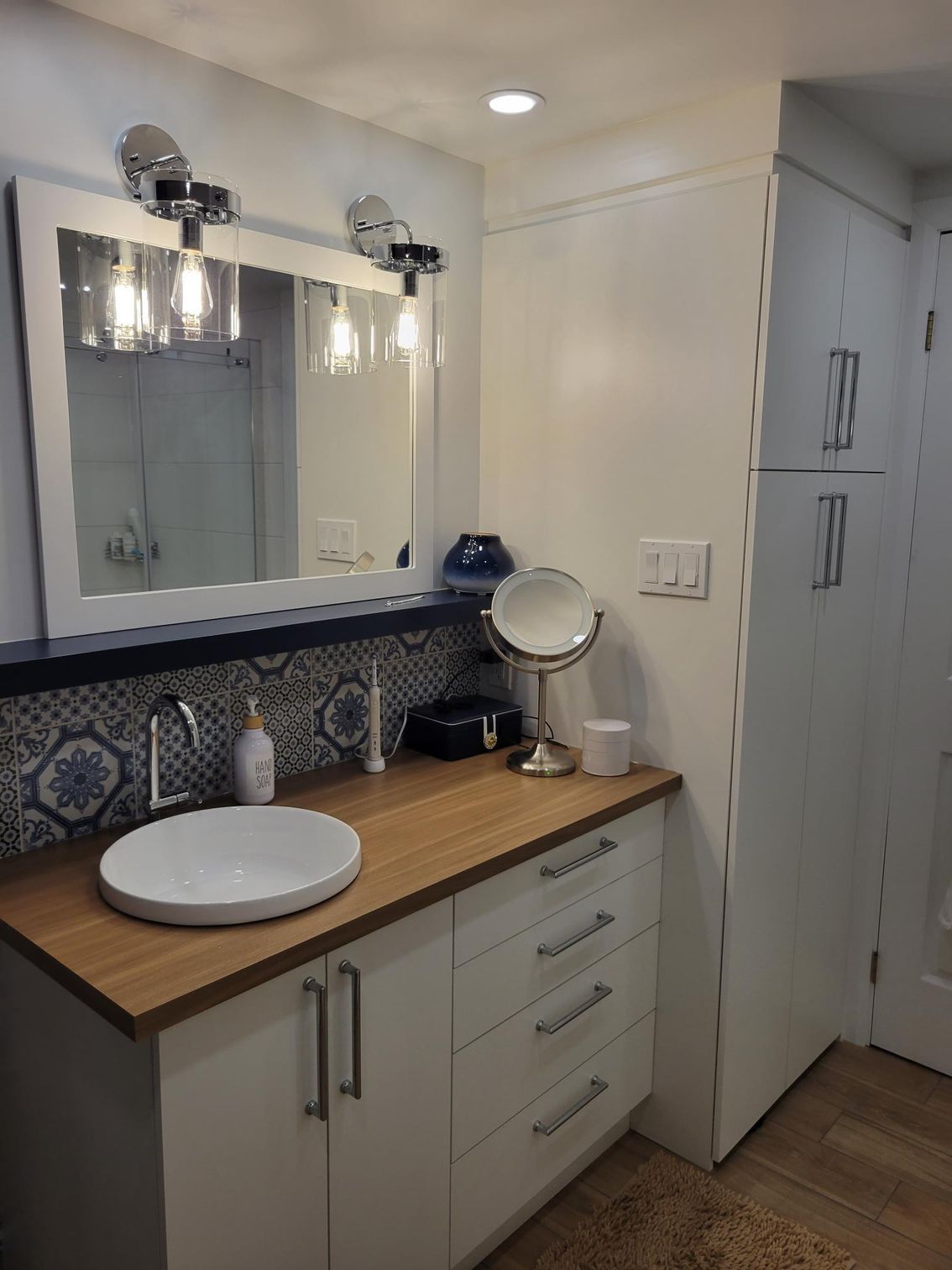 Salle de bain avec vanité blanche, comptoir en bois, lavabo rond, dosseret en carrelage bleu et miroir à trois lumières.