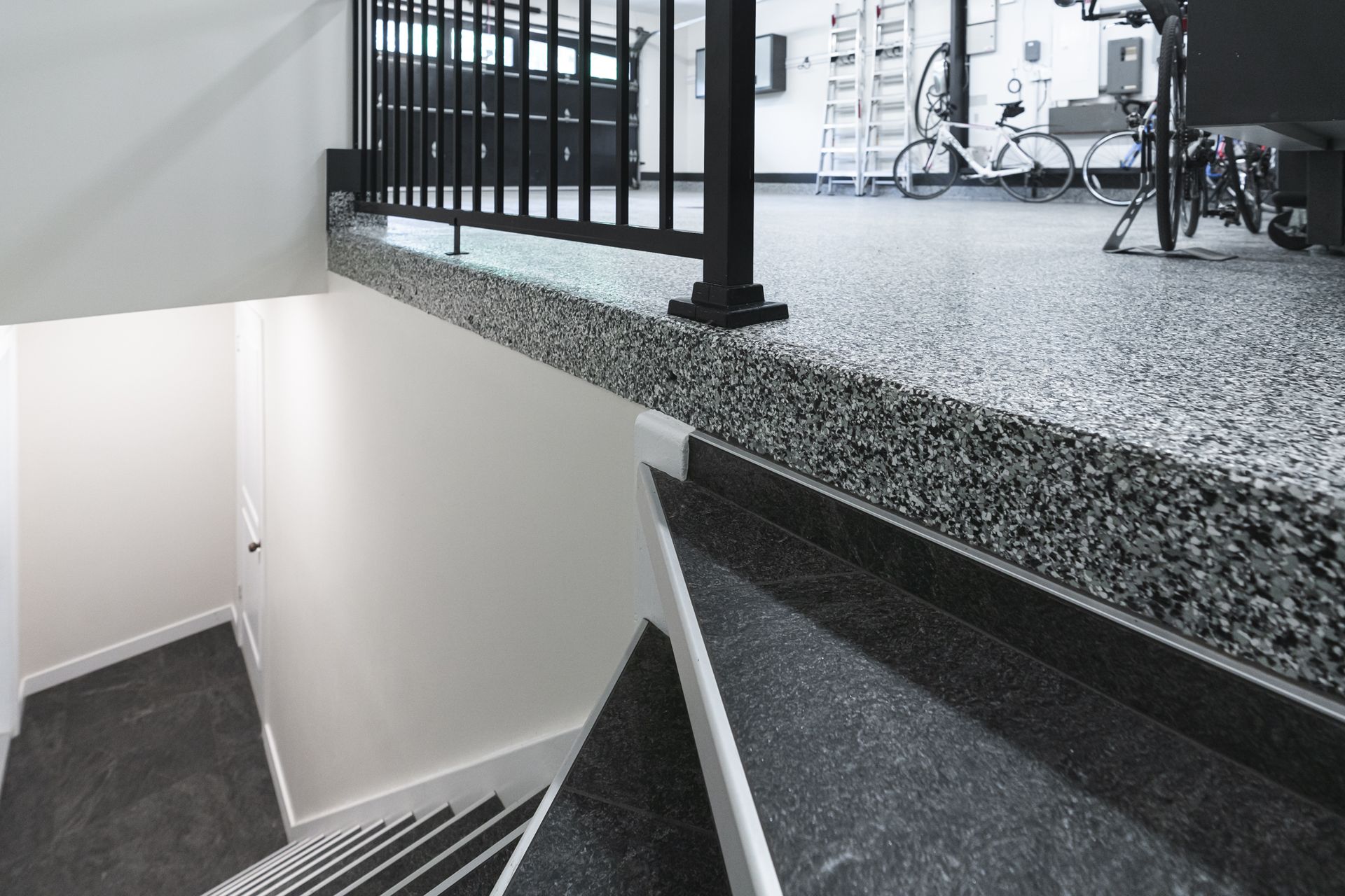 Un escalier descendant, vu depuis un balcon. Le balcon est doté d'une rampe noire et d'un sol gris moucheté.