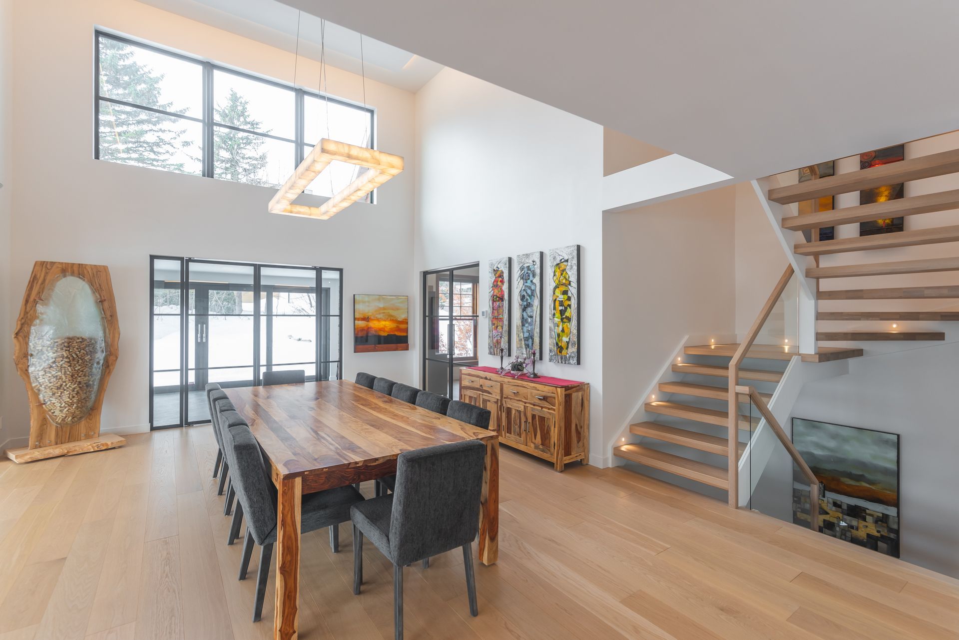 Salle à manger spacieuse avec une grande table en bois et un escalier, présentant de l'art moderne et de grandes fenêtres.