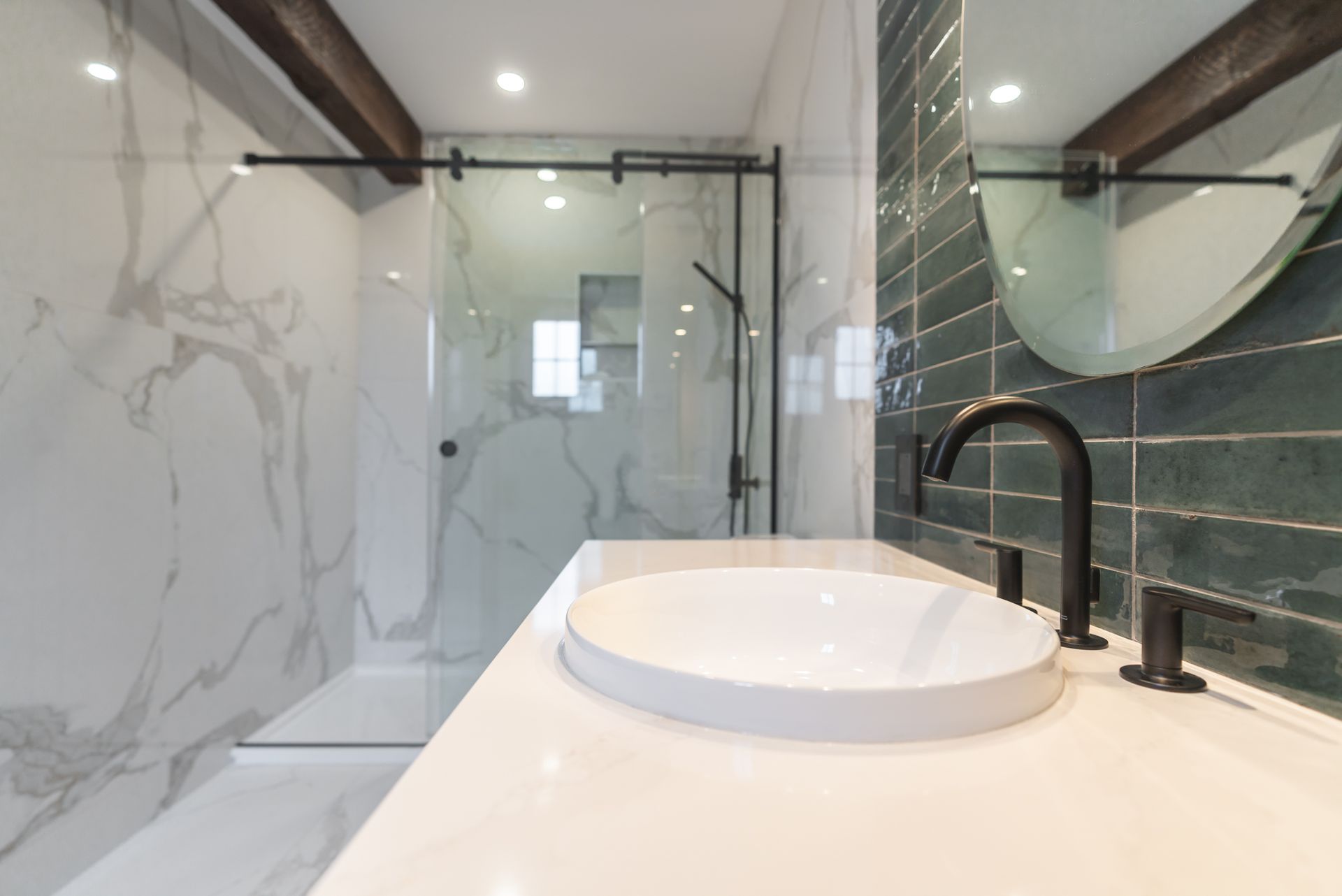 Salle de bain moderne avec comptoir blanc, lavabo rond, robinet noir, douche en verre et carrelage vert.