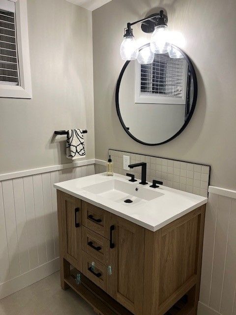 Une salle de bain lumineuse avec une vanité en bois, un miroir rond et des luminaires noirs contre des murs neutres.