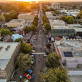 Aerial view of a city street lined with trees and low-rise buildings at sunset, with cars and pedestrians below