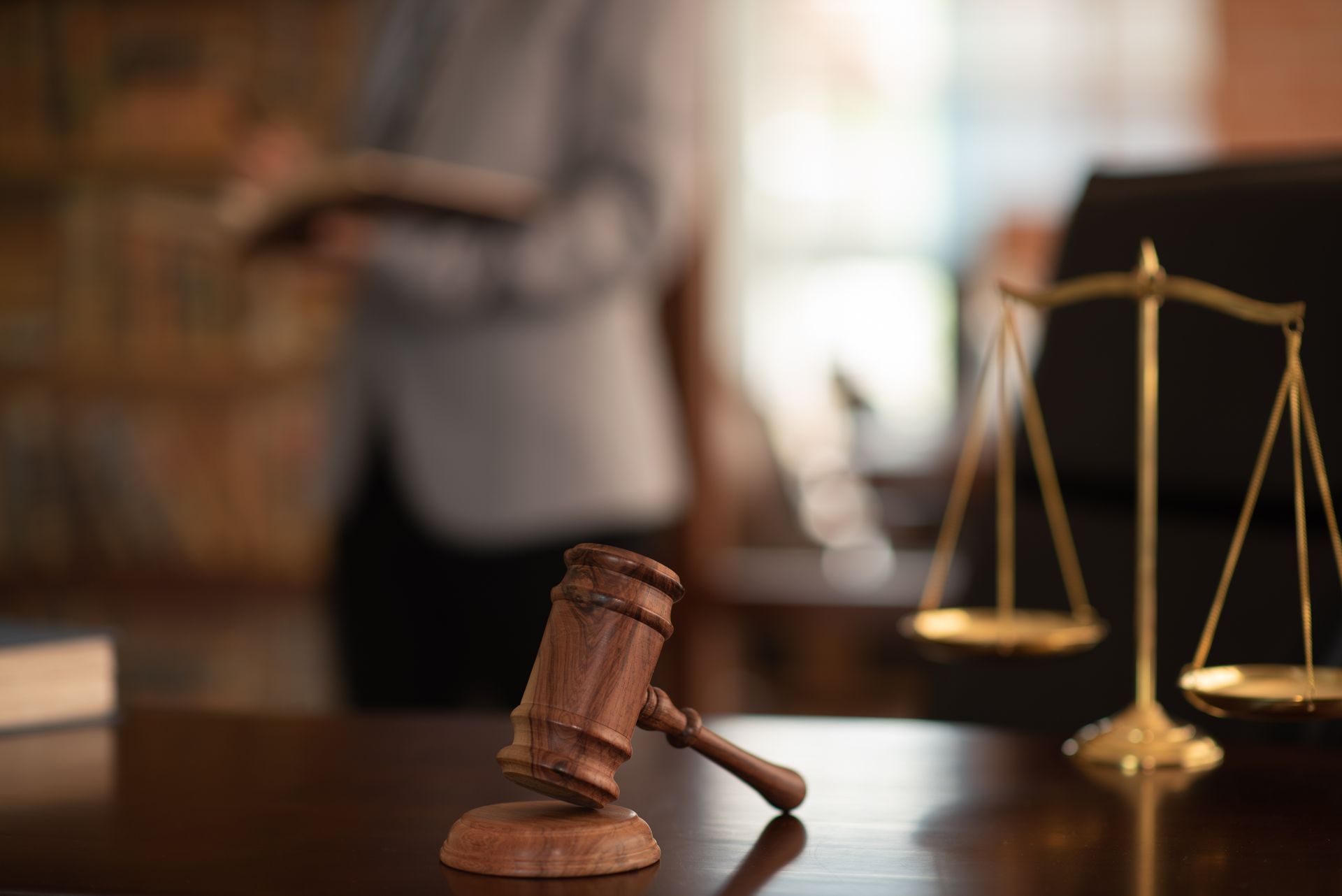 Gavel and scales of justice on a desk, a person in a suit is blurred in the background.