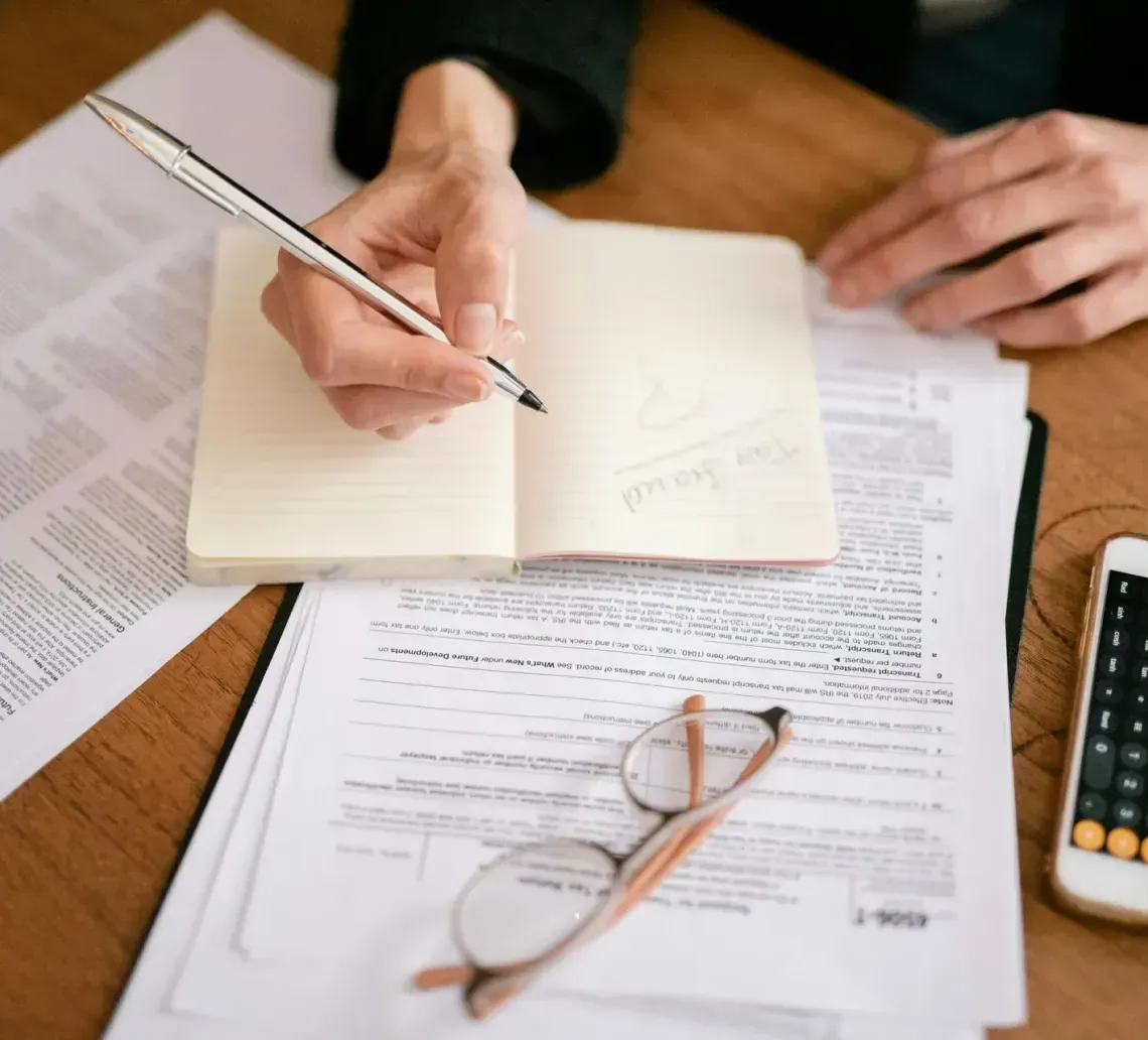 A person writing in a notebook on a wooden desk surrounded by tax forms, glasses, and a smartphone calculator.
