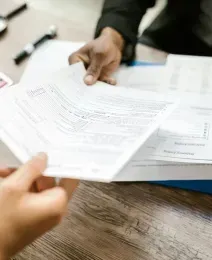 Two people exchange stacks of business documents over a wooden desk with a calculator and magnifying glass.
