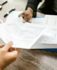 Two people exchange stacks of business documents over a wooden desk with a calculator and magnifying glass.
