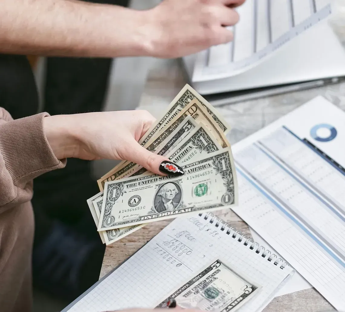 Two people managing finances, reviewing paper documents and counting US dollar bills while sitting at a table.