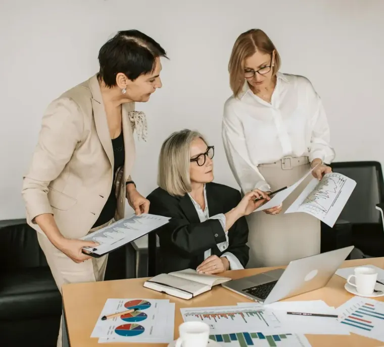 Three professionals in business attire collaborating over documents and charts on a desk in an office.