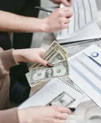 Two people managing finances, reviewing paper documents and counting US dollar bills while sitting at a table.