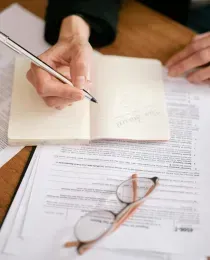 A person writing in a notebook on a wooden desk surrounded by tax forms, glasses, and a smartphone calculator.