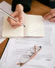 A person writing in a notebook on a wooden desk surrounded by tax forms, glasses, and a smartphone calculator.