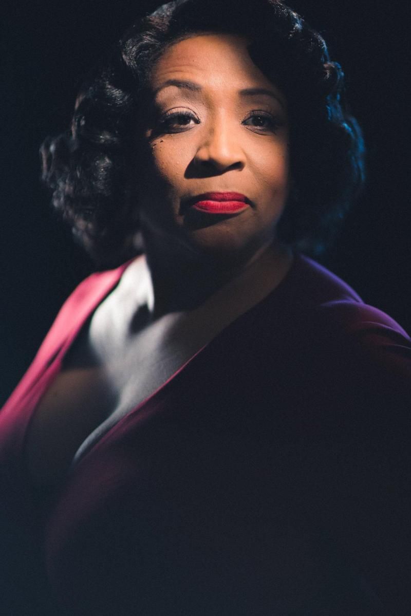 Woman in a red dress with red lipstick, dark hair, and a serious expression, lit by dramatic lighting.