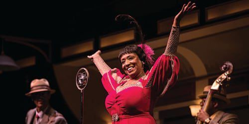 A woman in a red dress sings onstage, arms raised. A jazz band plays in the background.