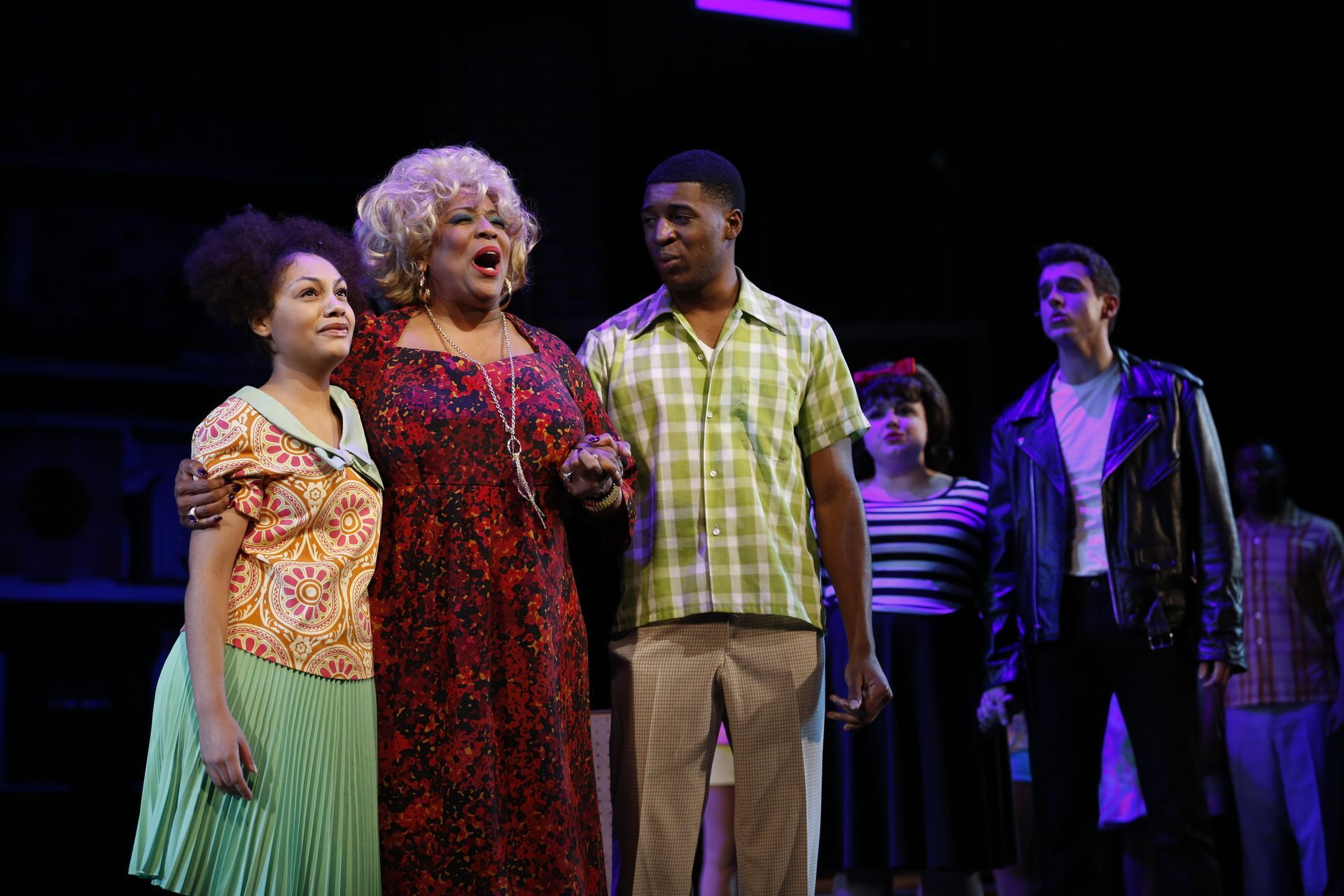 Stage cast in costume, smiling. Woman with curly blonde hair stands center with other performers.