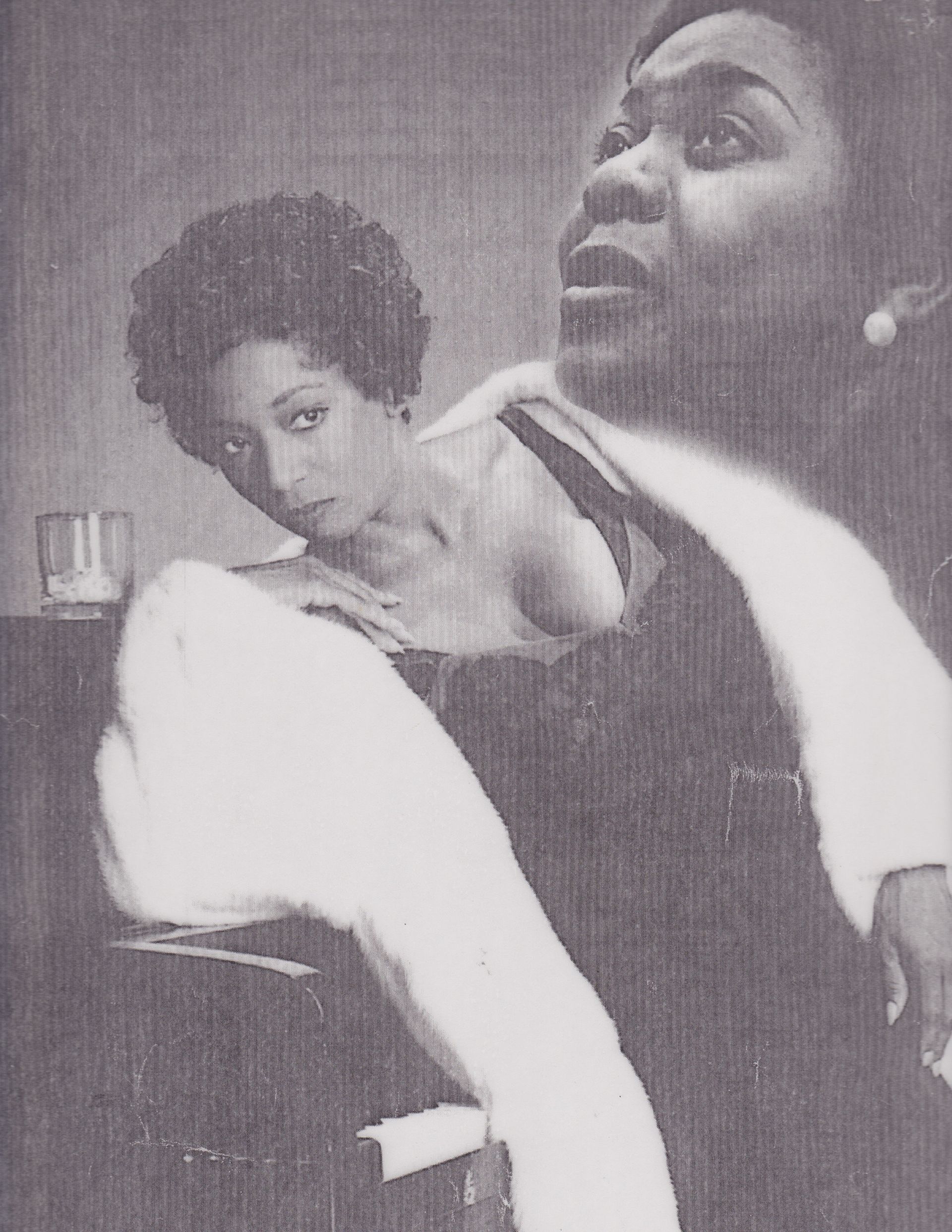Two Black women: one leaning on a piano, one singing, with a glass nearby. Black and white photo.