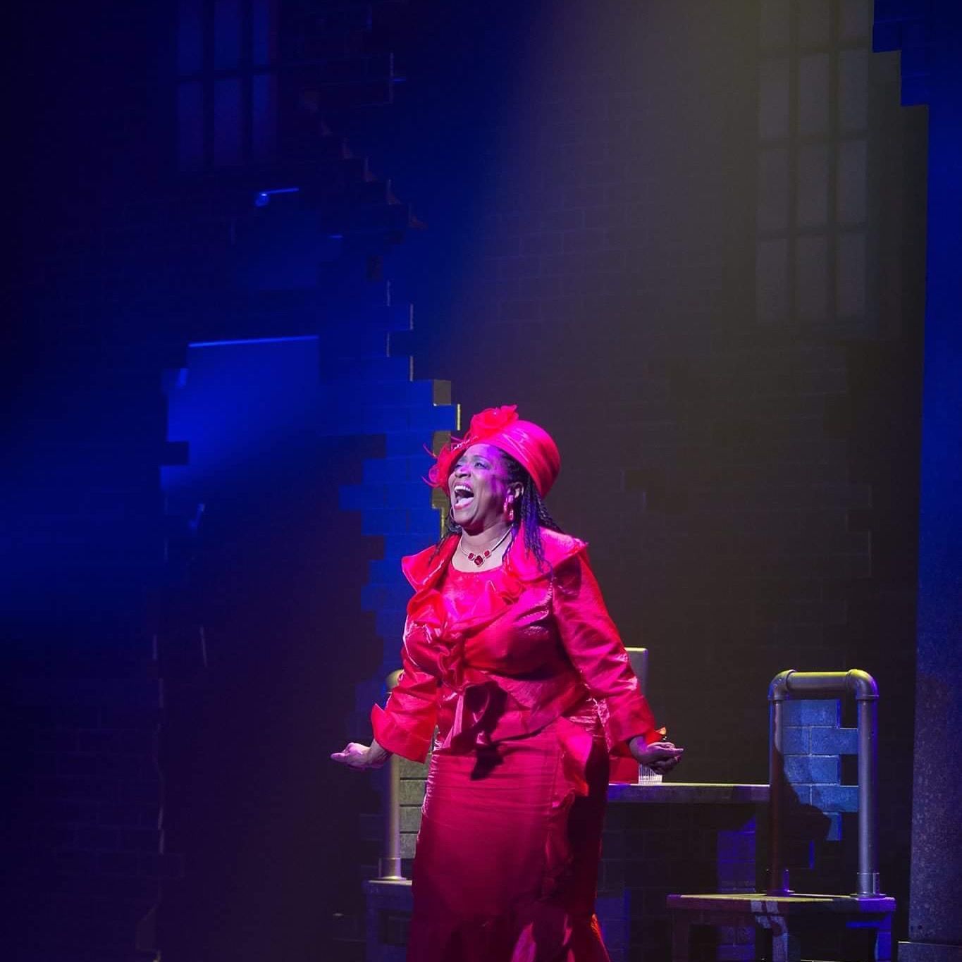 Woman in a red dress singing on a stage, arms outstretched, with a dramatic expression.