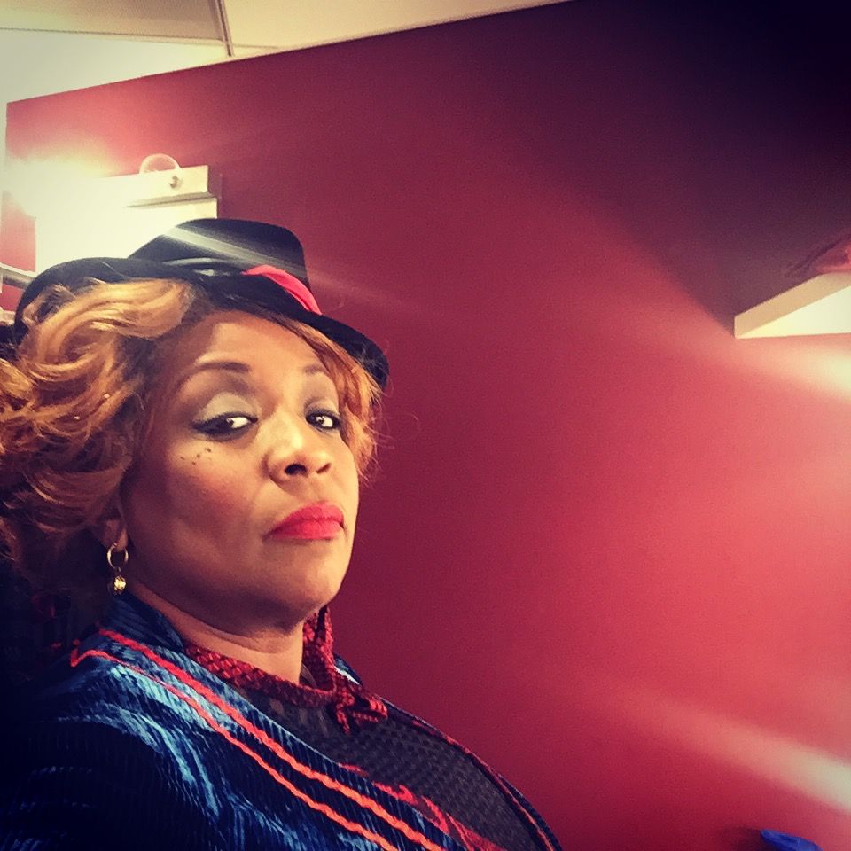 Woman with curly red hair, hat, red lipstick, in a red-walled room; looking at the camera.