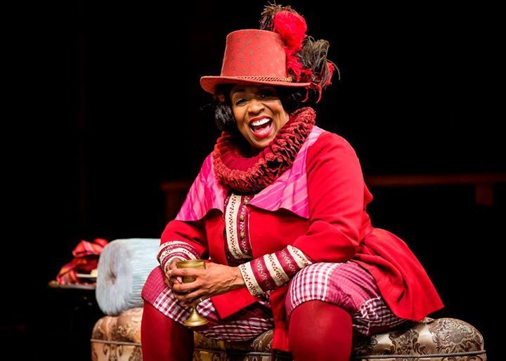 A Black woman in red period costume, smiling widely, sitting on a trunk on stage.