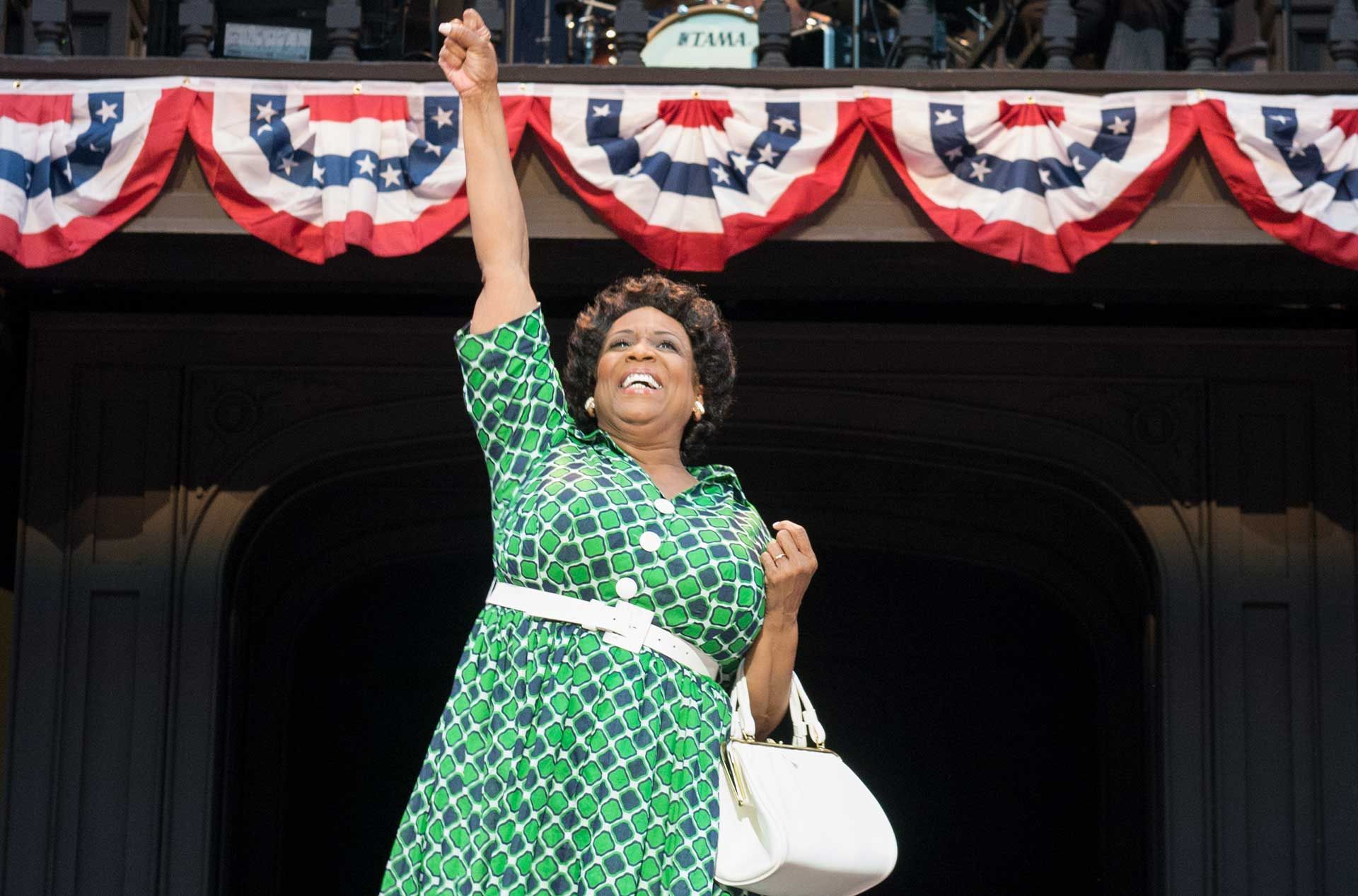 Woman in green dress raises arm, smiles on balcony; red, white, blue bunting overhead; white purse.