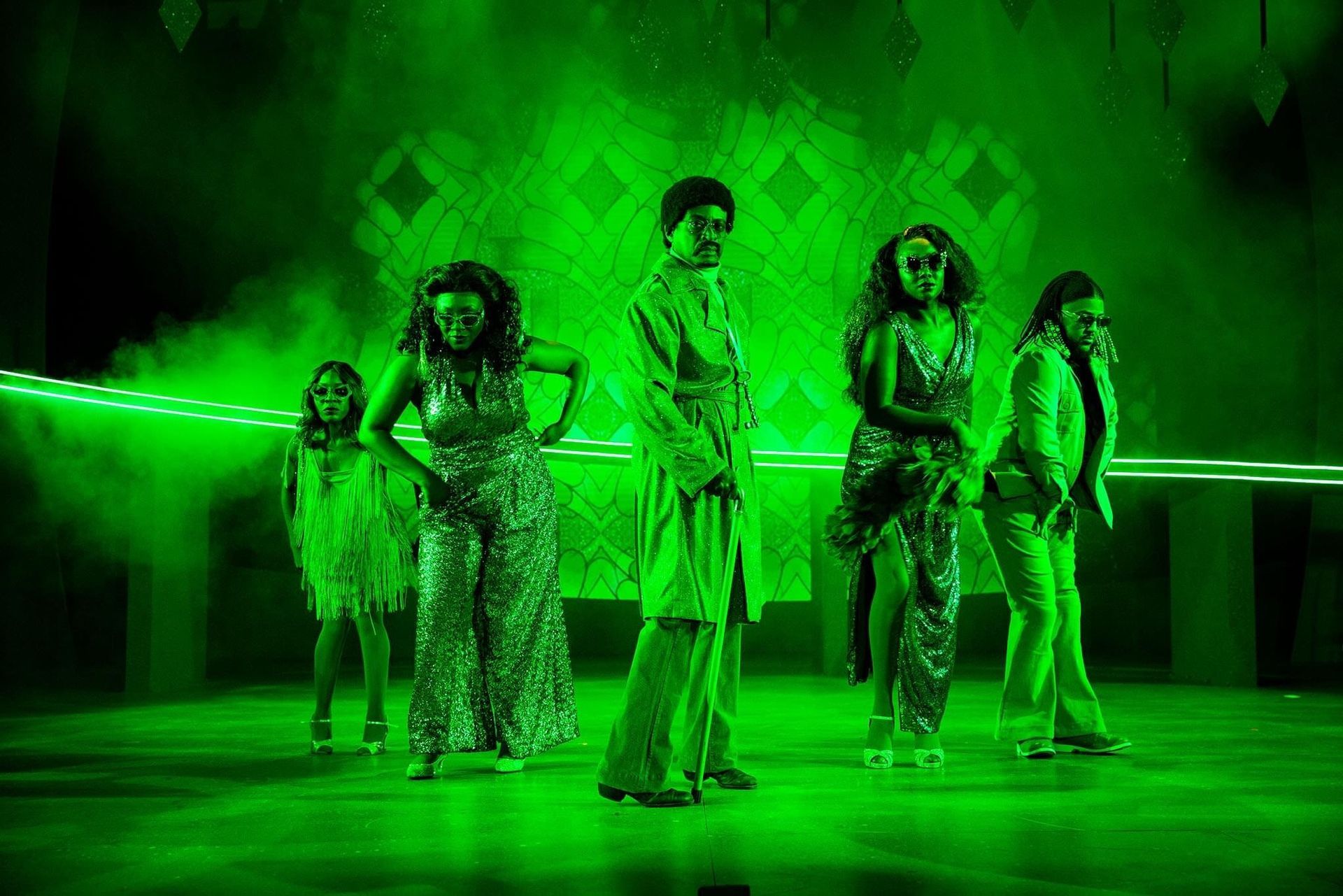 Five people in sparkly green outfits, on stage, bathed in green light, striking poses.