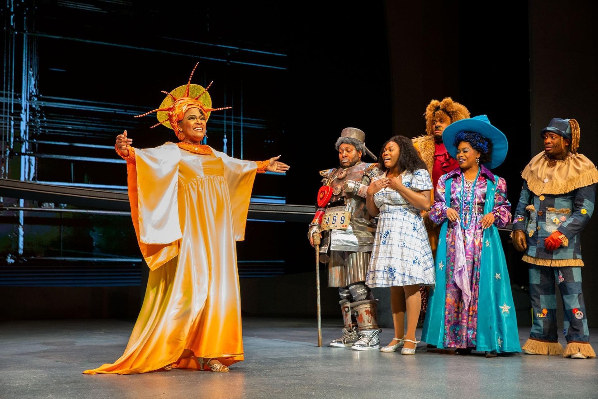 A stage performance of The Wizard of Oz, with a dressed character in gold, gesturing to the other characters.