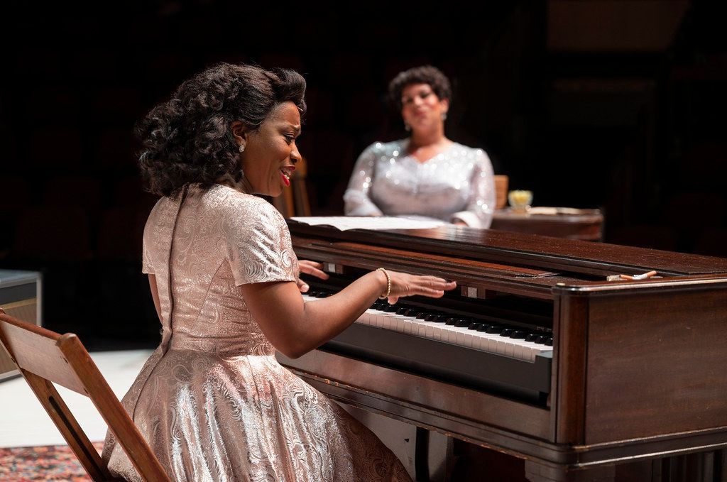 Woman in dress plays piano; another woman watches from behind. Stage.