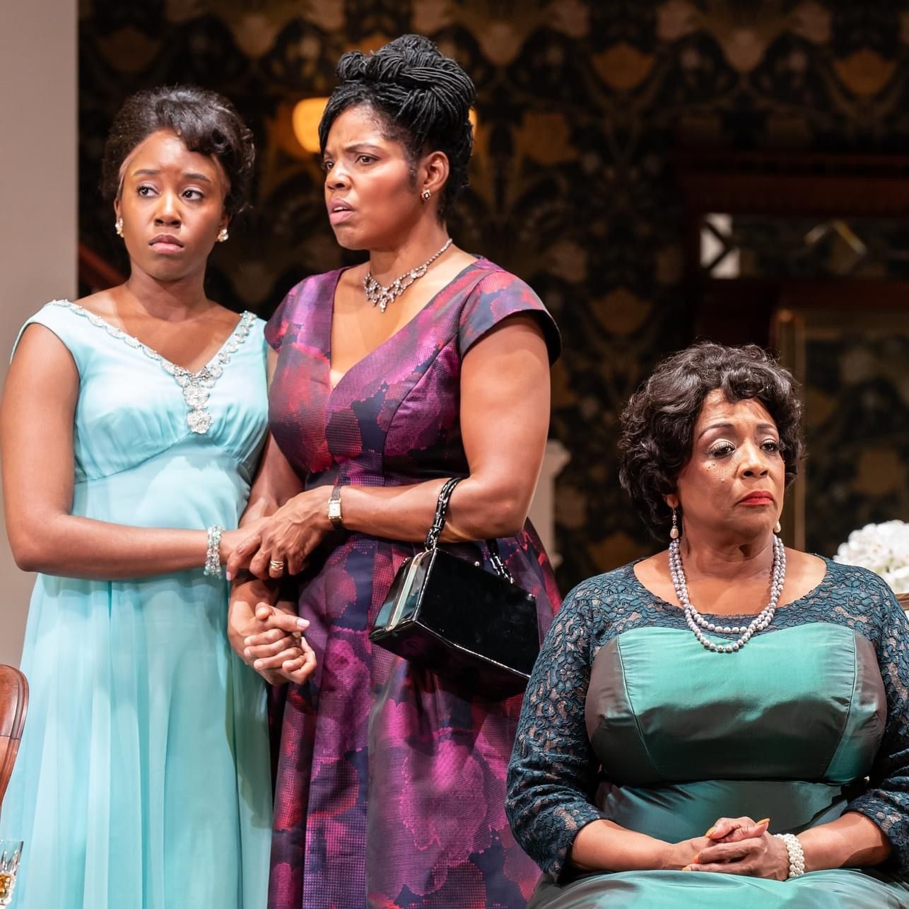Three Black women in a room; dressed in formal dresses, with concerned expressions.