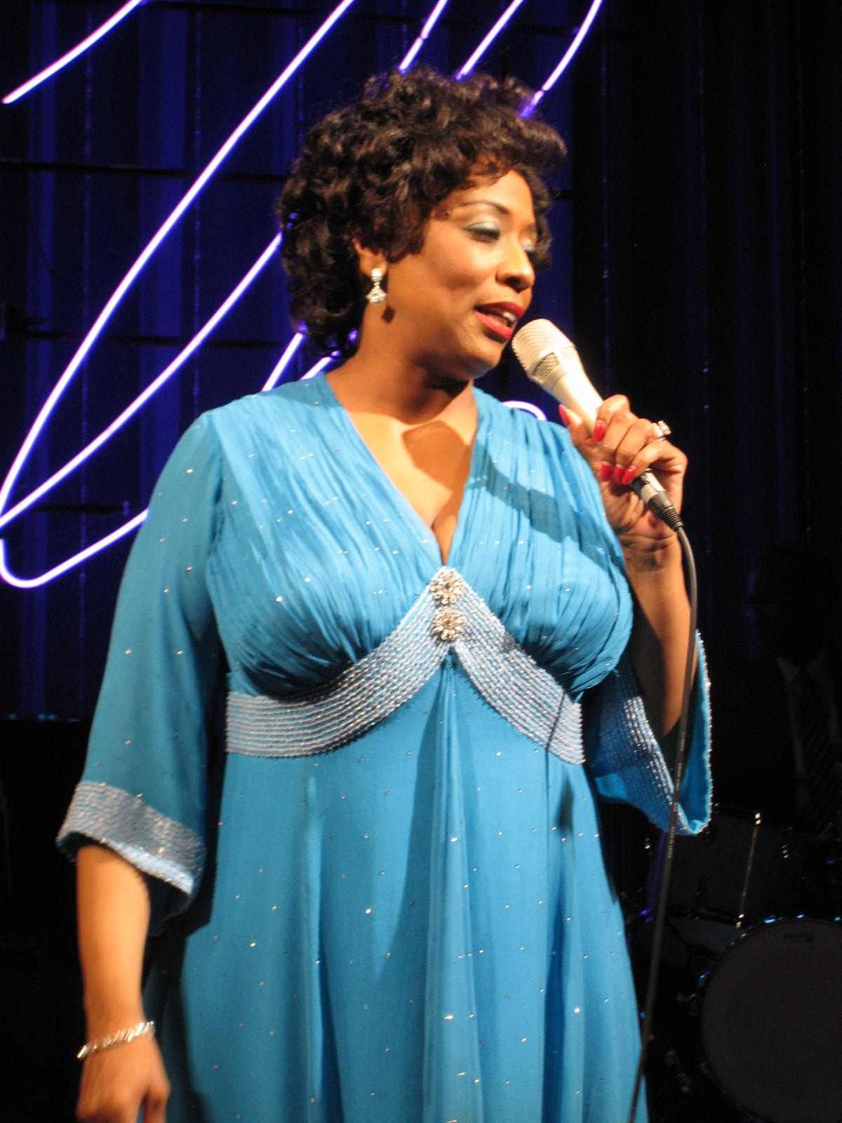 Woman in a blue gown singing into a microphone, stage with neon lights.