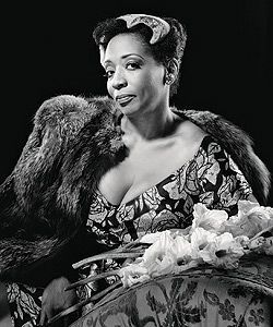 Woman in elegant dress and fur, seated, holding flowers; black and white portrait.