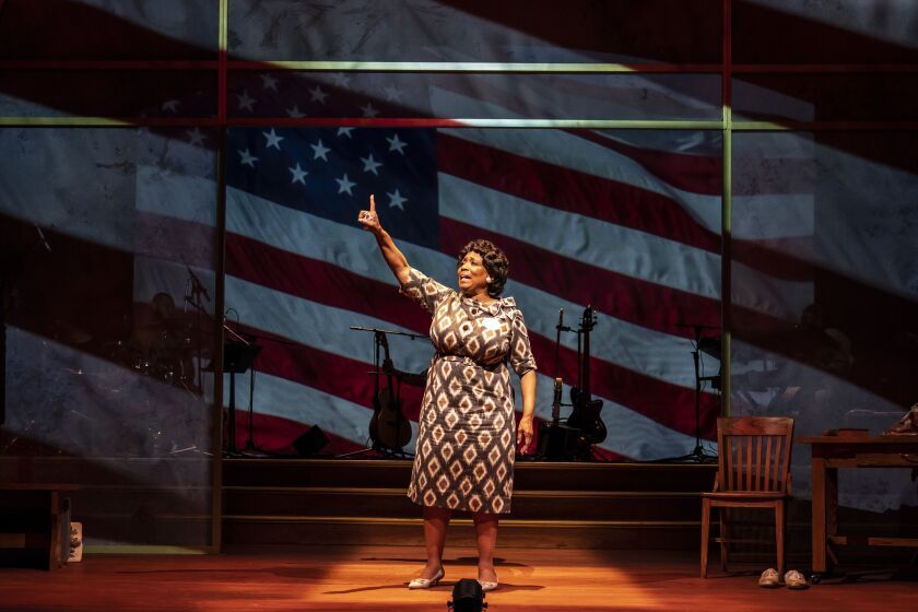 Woman on stage, pointing upwards in front of an American flag backdrop.