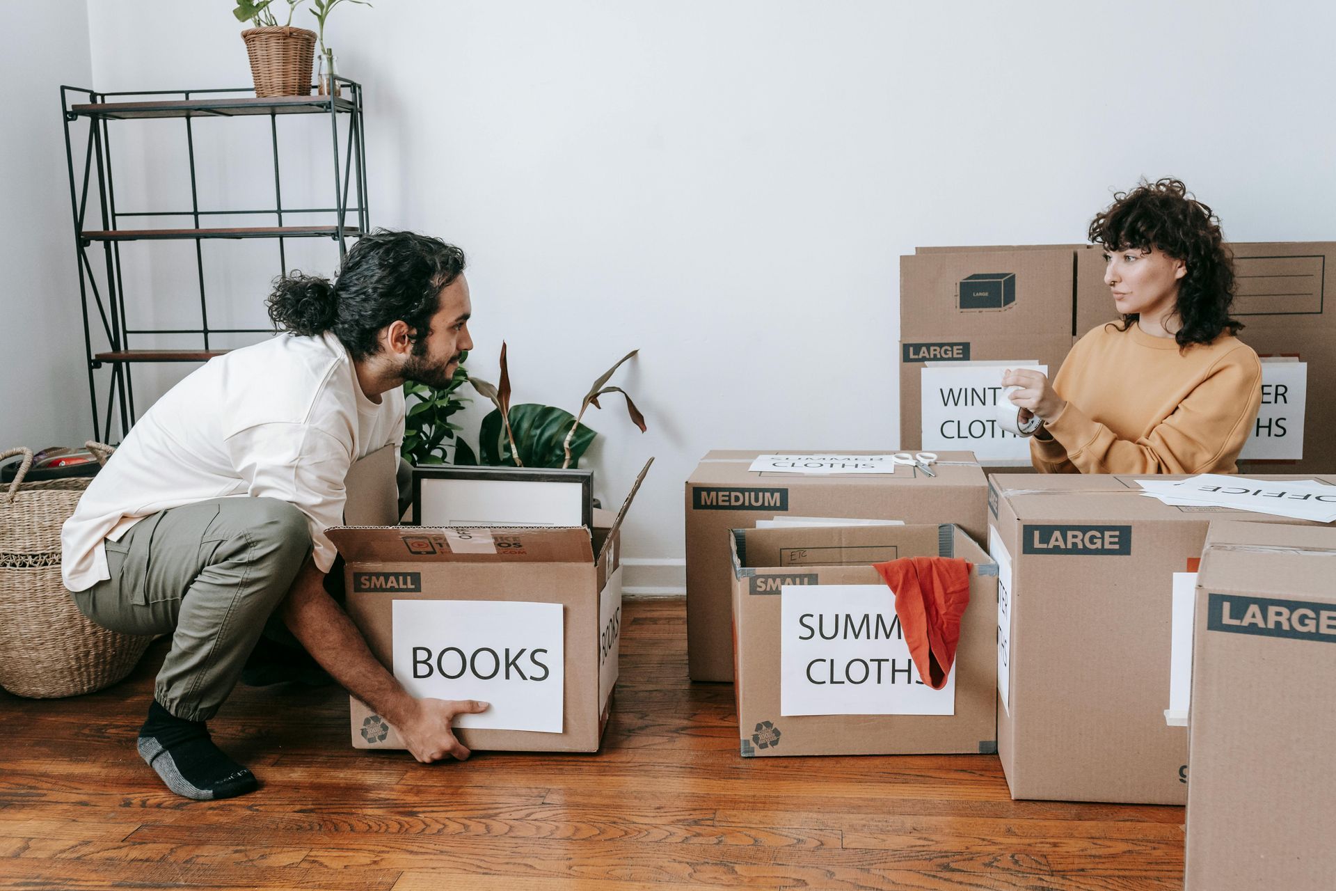 Two people packing moving boxes in a living room, with labeled boxes and a seated person smiling