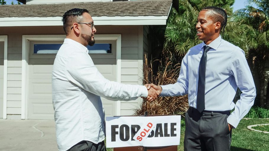 Two men shake hands in front of a house with a “For Sale” sign.