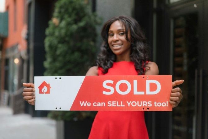 Woman in red dress holding a “SOLD” real estate sign on a city street