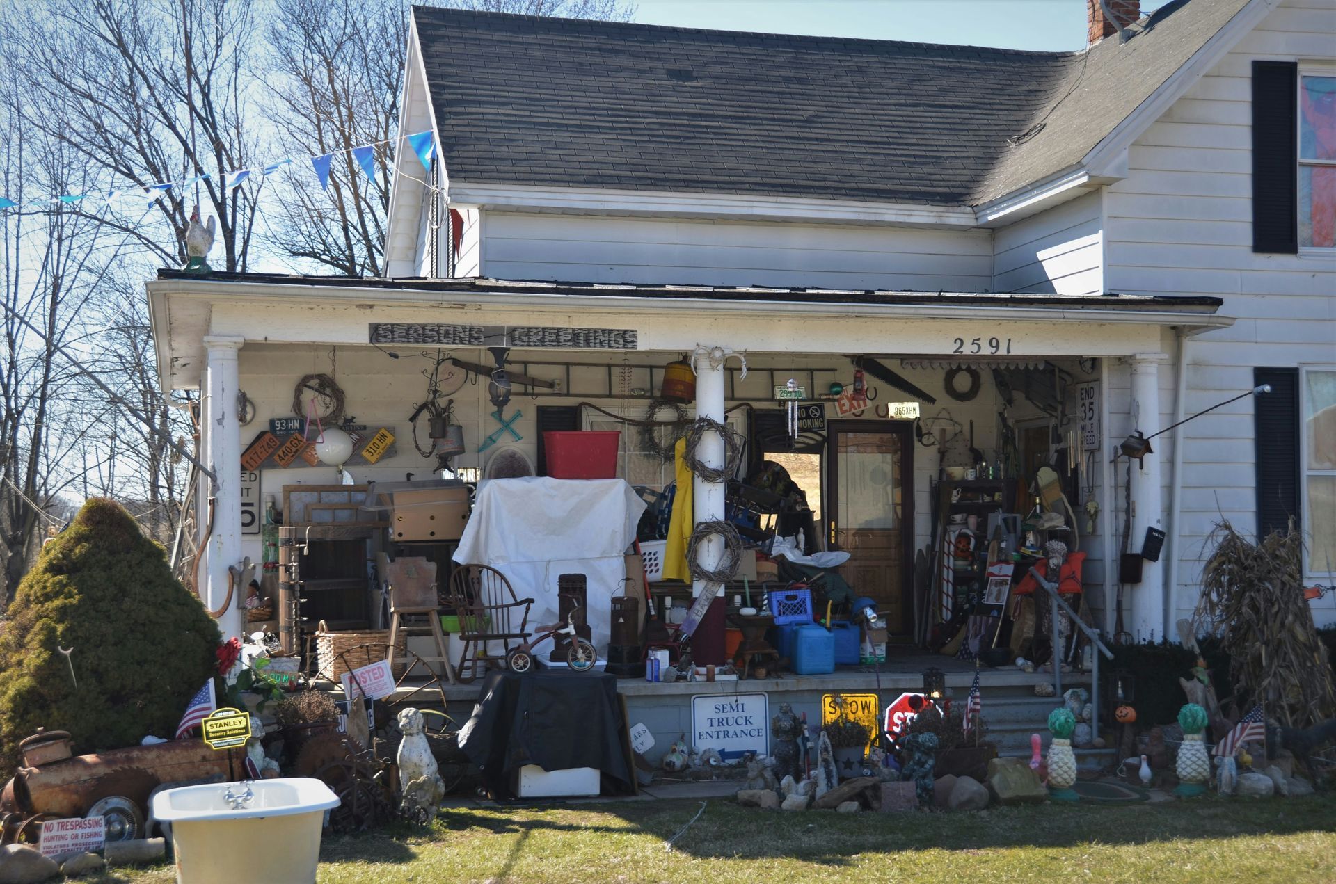 Cluttered front porch filled with tools, decorations, and household items on a suburban house.