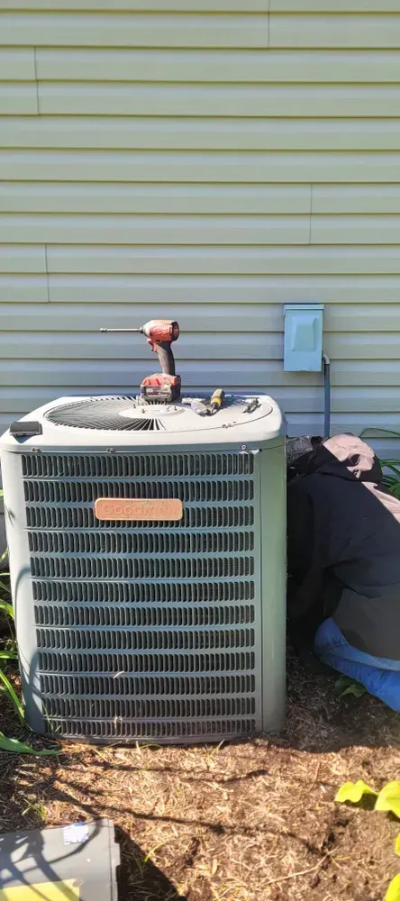 A person working on an outdoor air conditioning unit next to a house with tan siding.