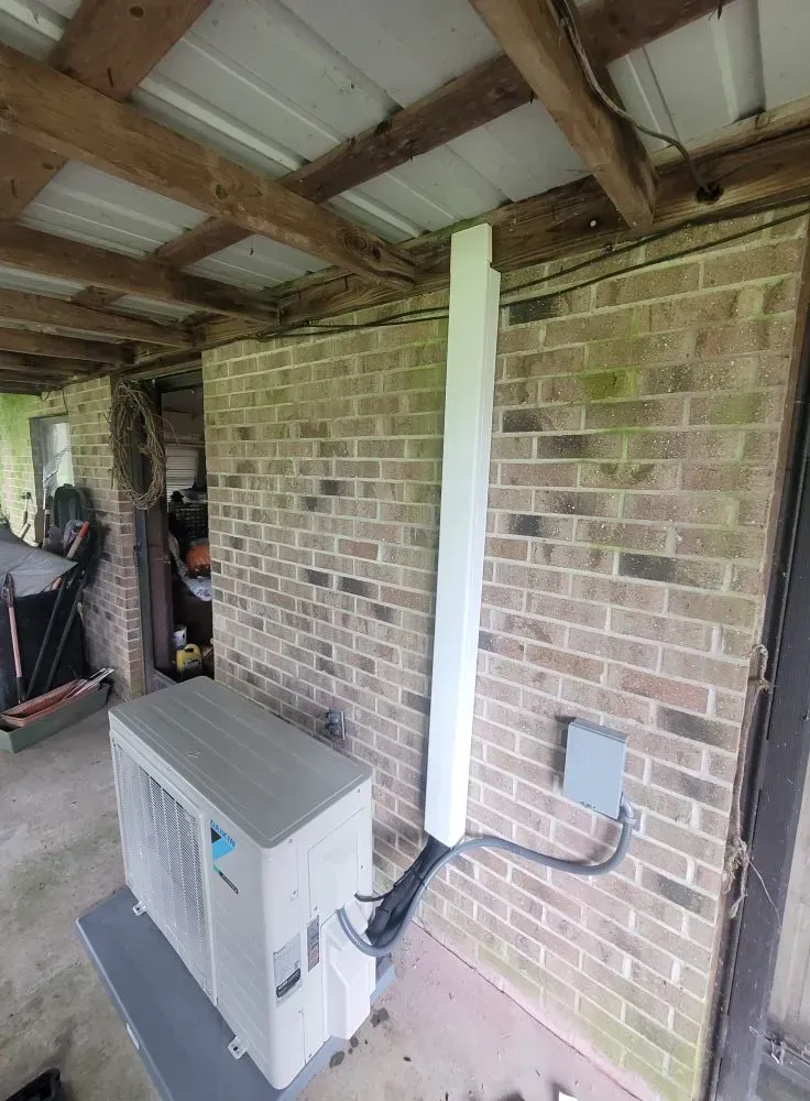 An outdoor HVAC unit next to a brick wall with a white conduit running up to the ceiling.