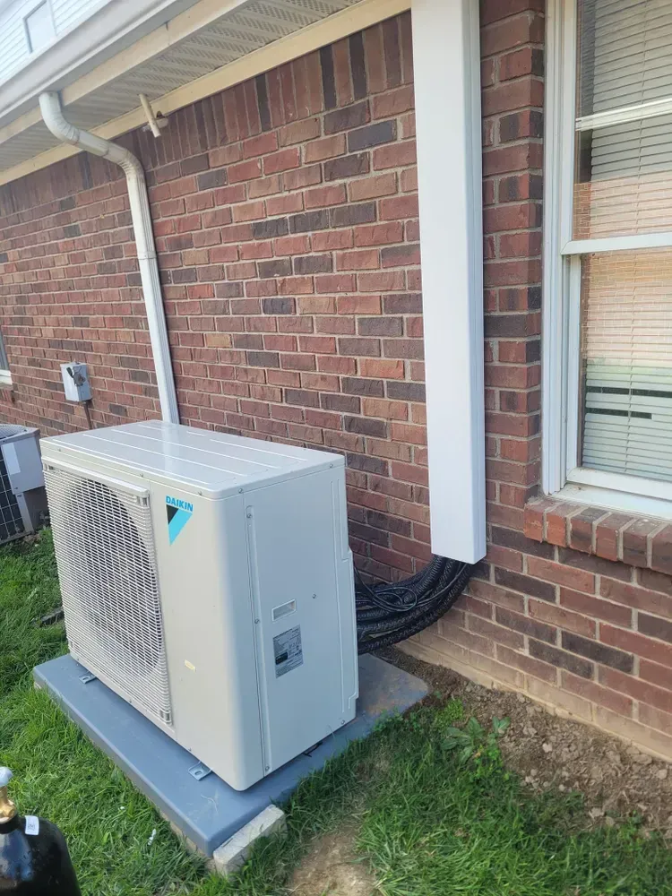 An air conditioning unit next to a brick building with a white conduit and a window.