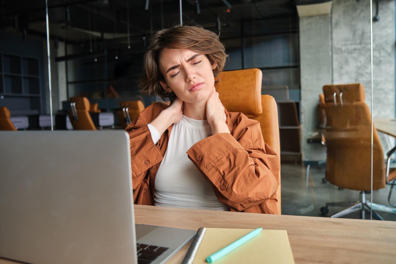 Person in an orange blazer massaging their neck while working on a laptop in an office