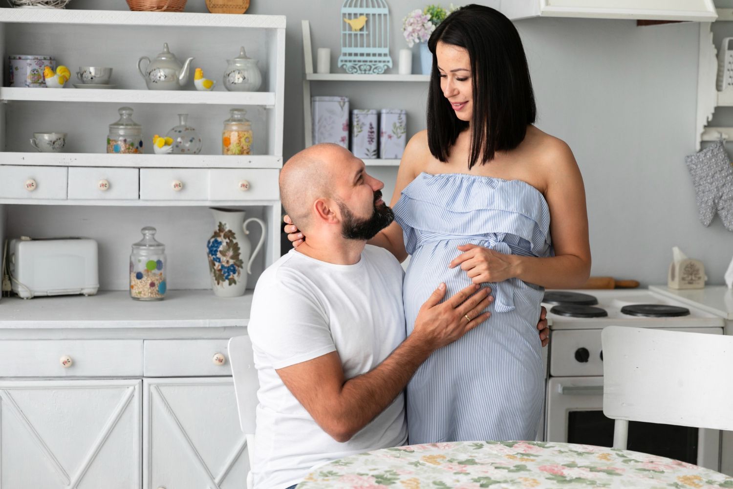 Man kneels and smiles while holding a pregnant person's belly in a bright kitchen