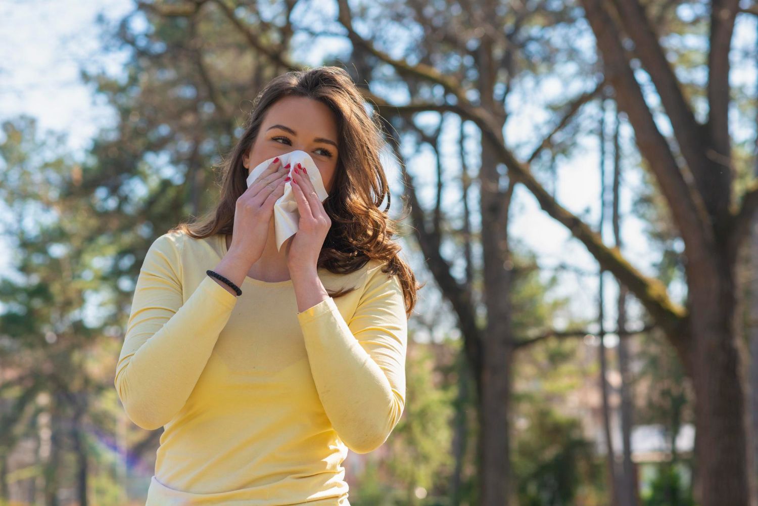 Woman outdoors in a yellow sweater, holding a tissue to her nose in a sunny park