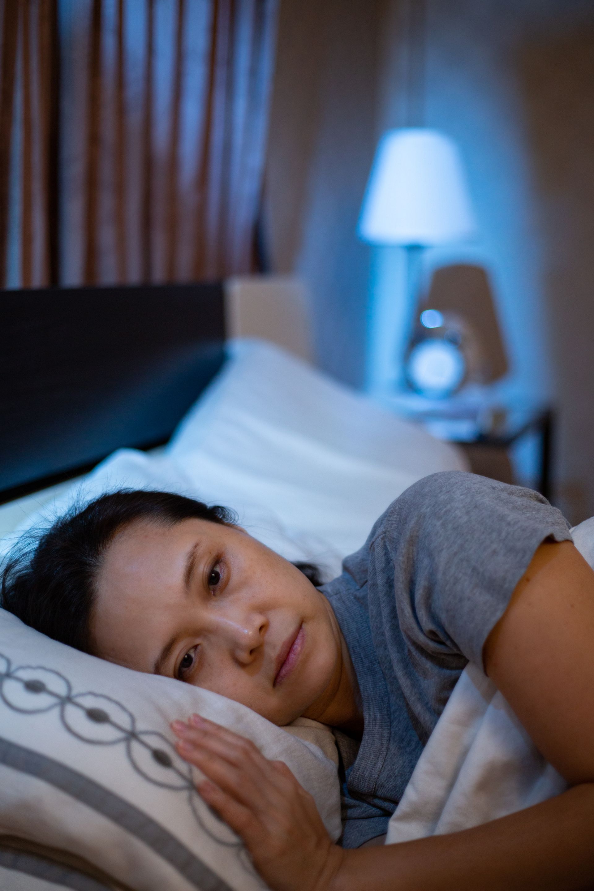 Person lying in bed, smiling softly under white blankets in a dim bedroom with a lamp glowing nearby