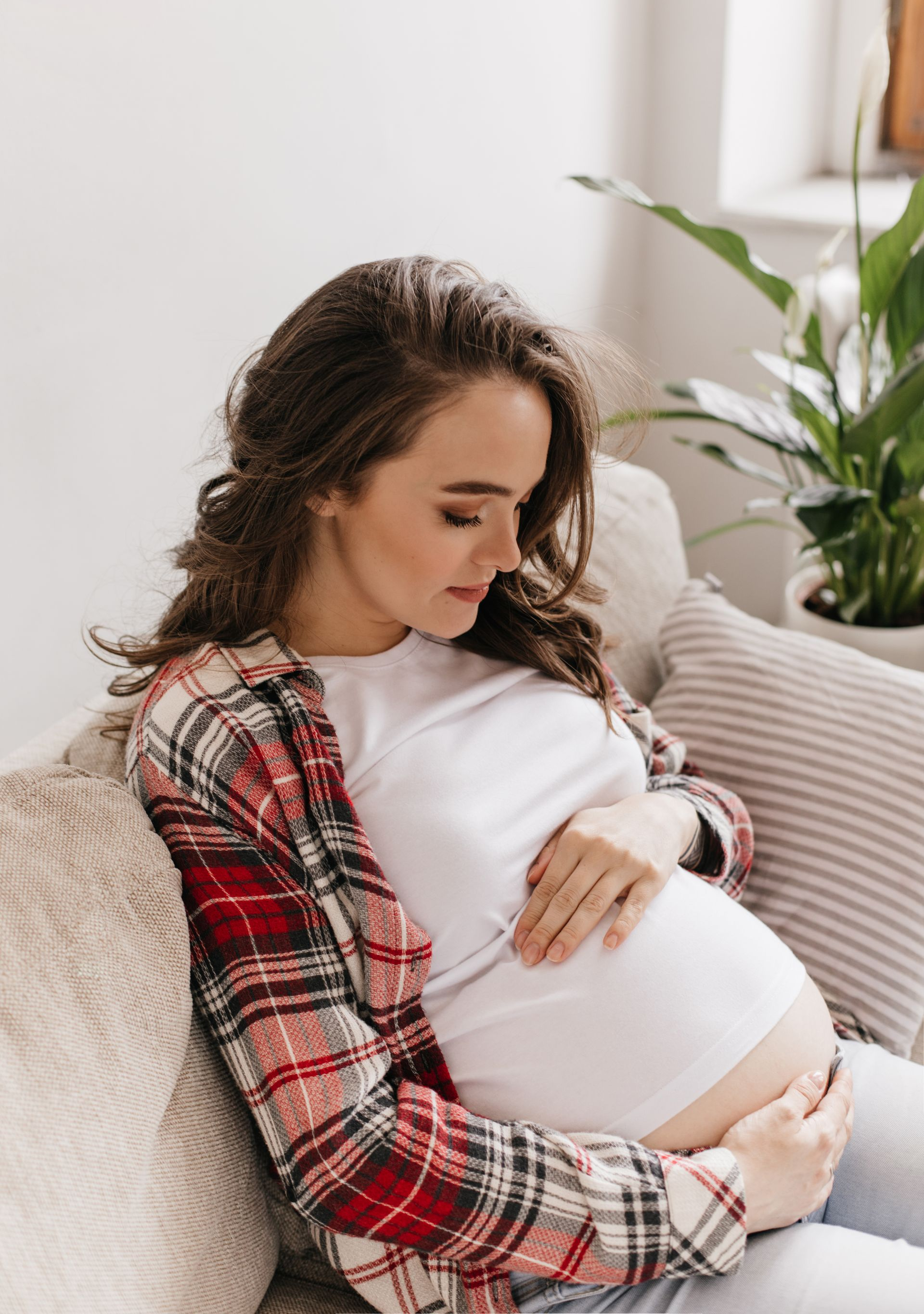 Pregnant person sitting on a couch, gently holding their belly by a bright window
