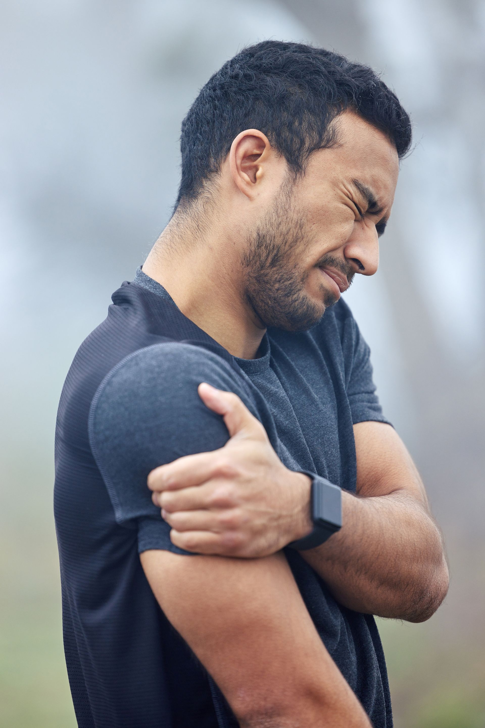 Man clutching his upper arm and grimacing in pain, wearing a dark shirt outdoors.