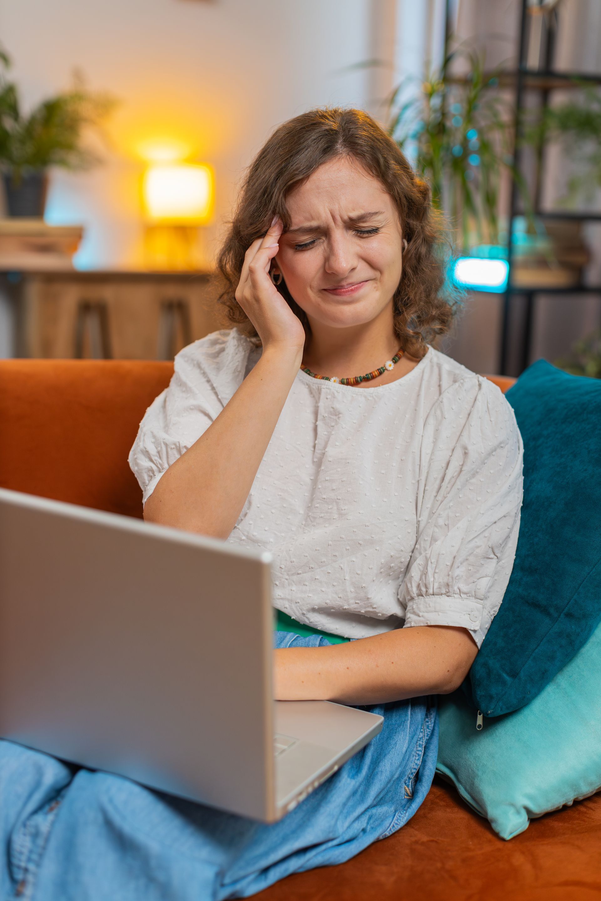 Woman on a couch with a laptop, holding her head and looking uncomfortable or tired.