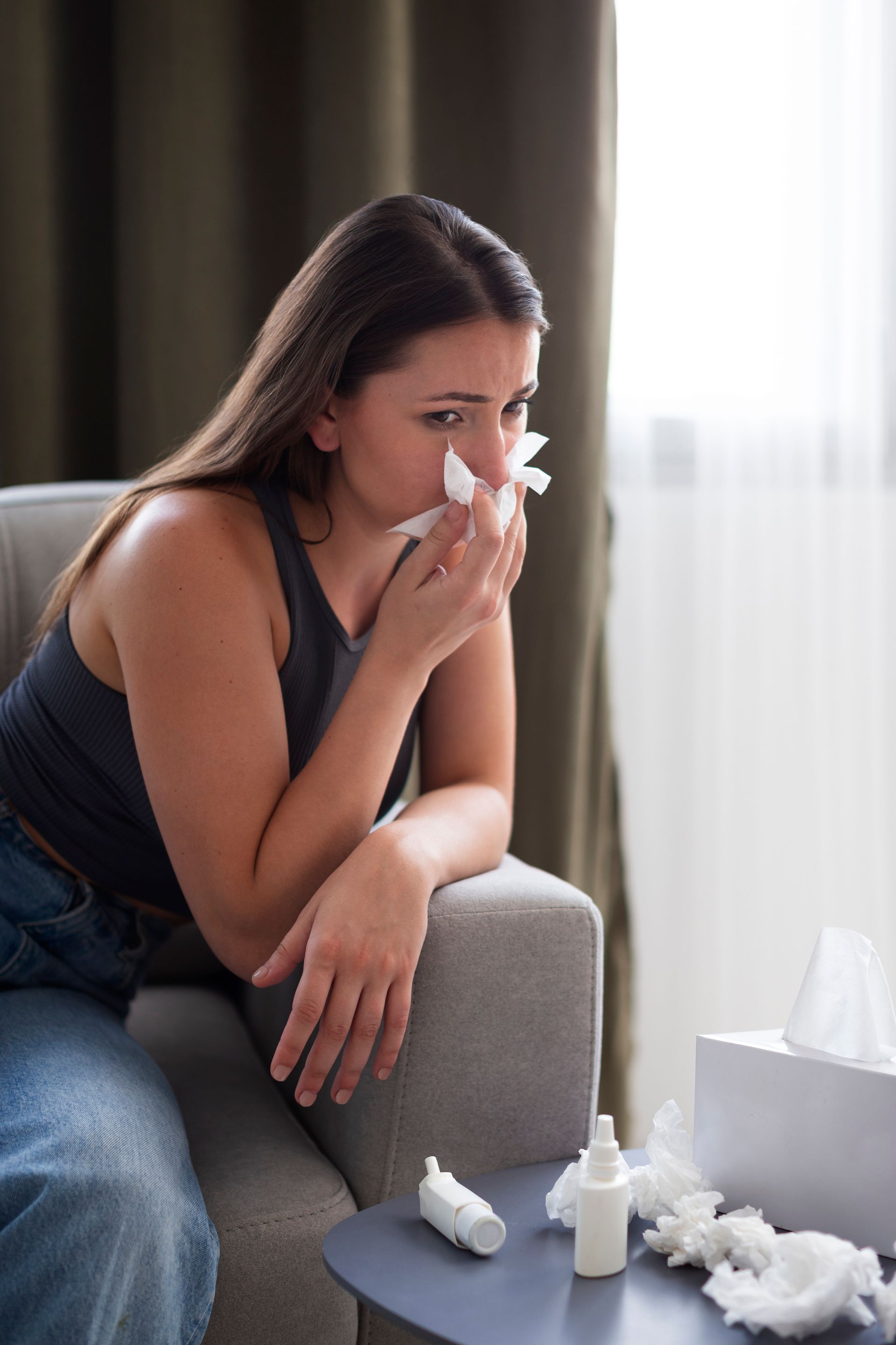 Woman sitting on a couch, blowing her nose with tissue beside a box of tissues and medicine bottles.