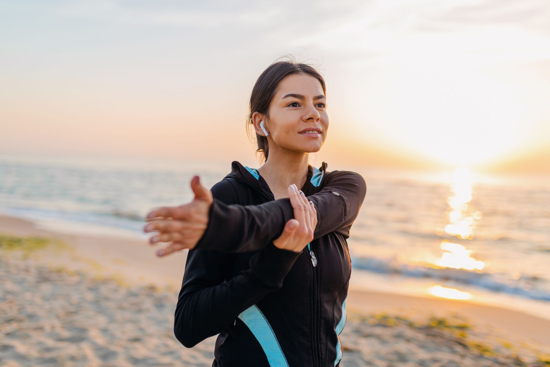 Woman stretching on a beach at sunrise, wearing a black and blue jacket.