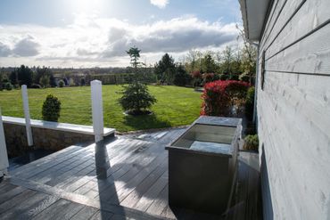 Backyard deck with wet boards, white posts, a green lawn, and a flower bed beside a house.