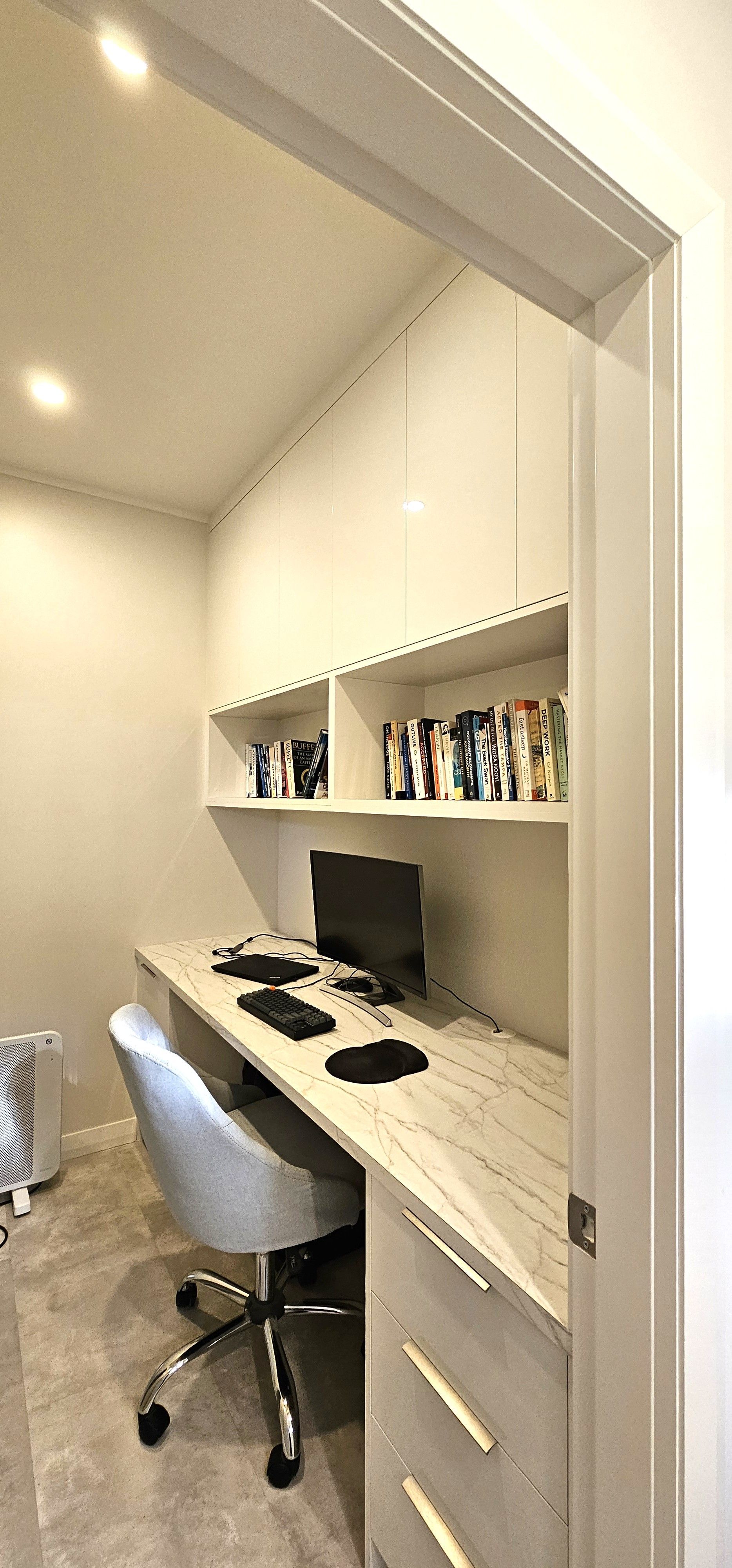 A home office with a built-in desk and overhead storage. A computer sits on the desk with a gray chair in front of it. The room is lit by overhead lights.
