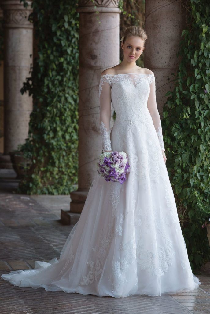 Eine Frau in einem weißen Hochzeitskleid hält einen Strauß lila Blumen in der Hand.